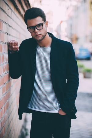 Young and handsome. Handsome young man in smart casual wear looking at camera while leaning at the brick wall with city view in the backgroundの写真素材
