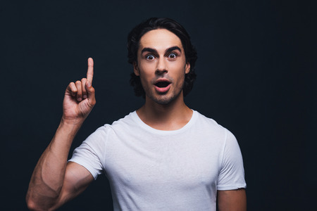 Did you see that? Portrait of surprised young man keeping mouth open and staring at camera while pointing up and standing against grey backgroundの写真素材