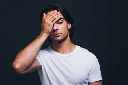 Feeling despair. Hopeless young man covering face with hand while standing against grey background
