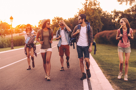 Enjoying summer hike. Group of young people with backpacks walking together by the road and looking happyの写真素材