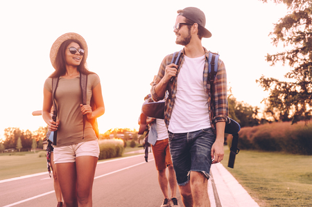 They love hiking. Group of young people with backpacks walking together by the road and looking happyの写真素材