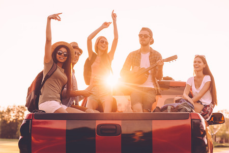 Enjoying road trip together. Group of young cheerful people enjoying their road trip while sitting in pick-up truck togetherの写真素材
