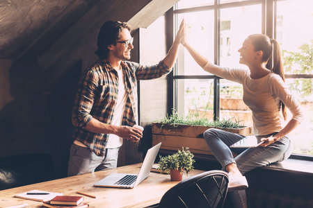 High-five! Cheerful young man and woman giving high-five while having coffee break in creative...
