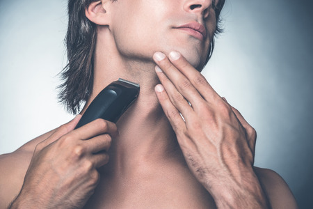 Shaving with electric razor. Close-up of handsome young shirtless man shaving with electric razor while standing against grey backgroundの写真素材