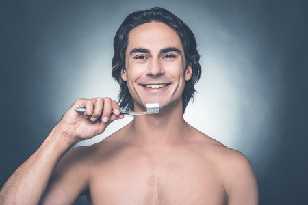 Man brushing teeth. Handsome young shirtless man brushing teeth and smiling while standing against grey backgroundの写真素材