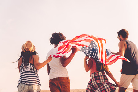 Friends with American flag. Rear view of four young people carrying american flag while running outdoorsの写真素材