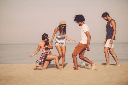 Beach ball with friends. Group of cheerful young people playing with soccer ball on the beach with sea in the backgroundの写真素材