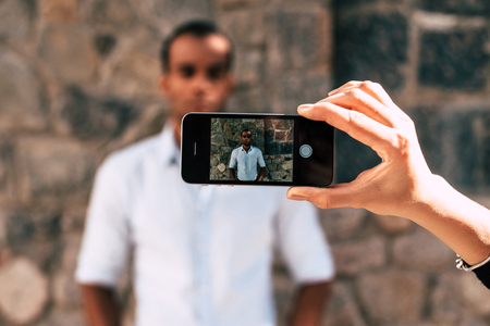 Taking a picture. Female hand holding smart phone and taking picture of young African man standing outdoorsの写真素材