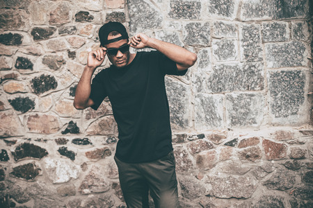 Confident in his style. Handsome young African man in casual clothes adjusting his sunglasses while standing against the stoned wall outdoorsの写真素材