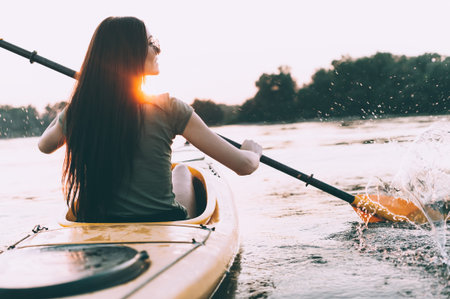 Enjoying perfect sunset on river. Rear view of beautiful young woman kayaking on river and with...