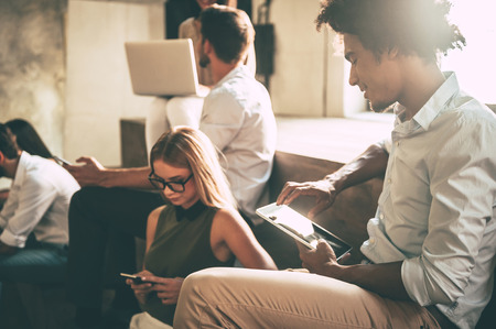 Living in a digital age. Group of young people using different gadgets while sitting close to each other indoorsの写真素材