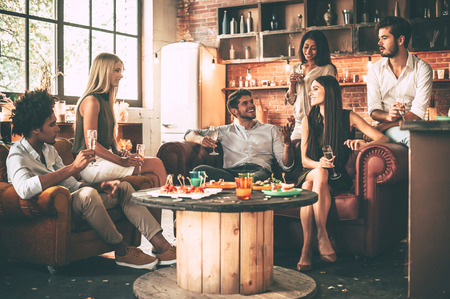 Enjoying time with best friends. Group of cheerful young people enjoying food and drinks while spending nice time in cofortable chairs on the kitchen togetherの写真素材