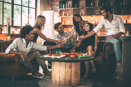 Cheers! Group of cheerful young people enjoying food and drinks while spending nice time in cofortable chairs on the kitchen togetherの写真素材