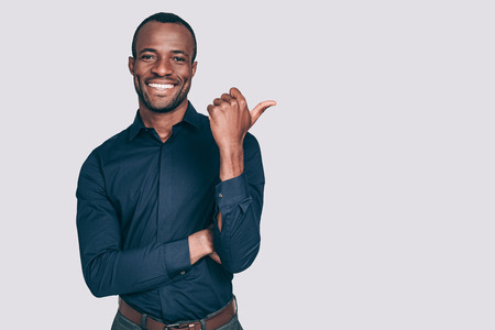 Look over there! Handsome young African man pointing away and smiling to you while standing against grey backgroundの写真素材