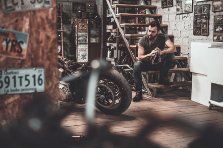 He needs a little break. Confident young man holding rag and looking at motorcycle while sitting near it in repair shopの写真素材