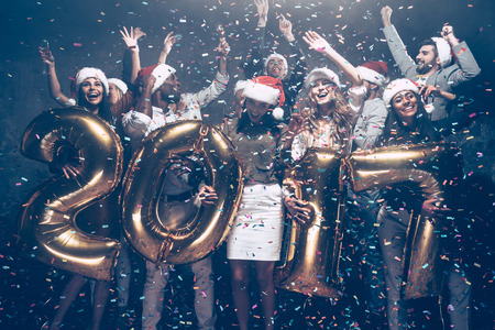 New Year is coming! Group of cheerful young people in Santa hats carrying gold colored numbers and throwing confettiの写真素材