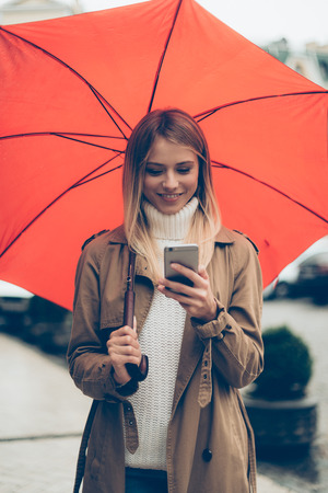 Quick message on the go. Attractive young smiling woman carrying umbrella and looking at her mobile phone while walking by the streetの写真素材