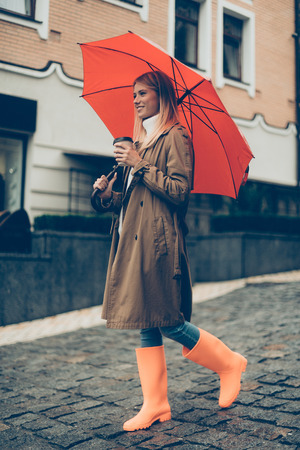 Enjoying autumn in city. Full length of attractive young smiling woman in rubber boots carrying umbrella and coffee cup while walking down the streetの写真素材