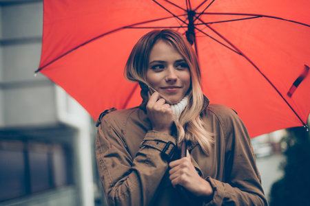 Feeling cold. Attractive young smiling woman carrying umbrella and adjusting her coat while walking...