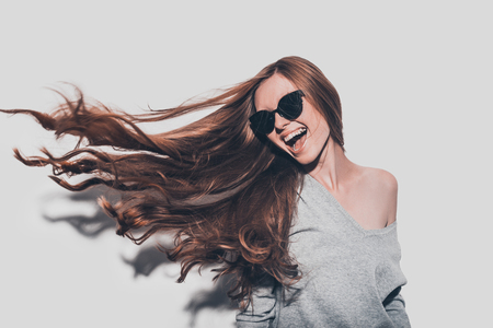 Hair like fire. Attractive young smiling woman in sunglasses and with tousled hair looking away while standing against grey backgroundの写真素材