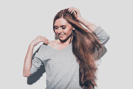 Cheerful beauty. Attractive young woman holding hand in hair and smiling while standing against grey backgroundの写真素材