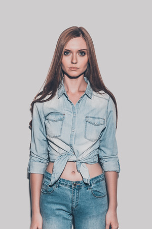 Jeans style. Studio shot of attractive young woman in jeans wear looking at camera while standing against grey backgroundの写真素材