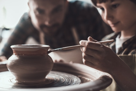 Pottery class. Little boy drawing on ceramic pot at the pottery class while man in apron standing close to himの写真素材