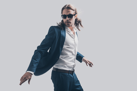 Style in motion. Handsome young man in full suit and sunglasses moving in front of grey backgroundの写真素材