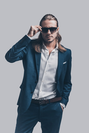 Used to look perfect. Handsome young man in full suit adjusting his sunglasses while standing against grey backgroundの写真素材