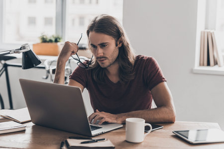 Full concentration at work. Confident young man with long hair carrying his eyeglasses and looking at laptop while sitting at his working place in officeの写真素材