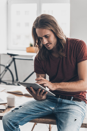 Modern businessman at work. Handsome young man with long hair working on digital tablet while sitting on the desk in creative officeの写真素材