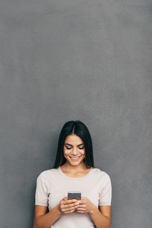 Texting to friend. Attractive young woman holding smart phone and looking at it with smile while standing against grey background with copy space upon her headの写真素材
