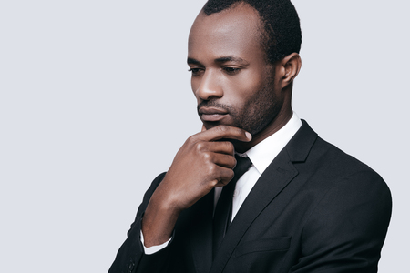 Difficult decision. Portrait of confident young African man holding hand on chin and looking away while standing against grey backgroundの写真素材