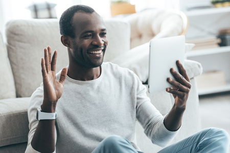 Hello! Handsome young African man looking at his digital tablet and smiling while sitting on the carpet at home の写真素材