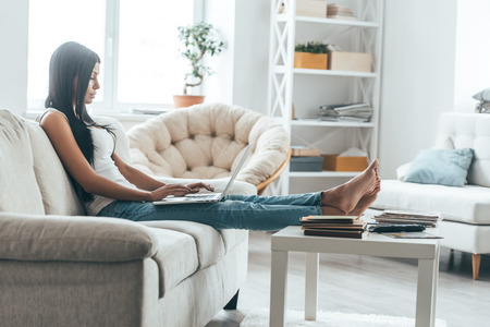 Writing lovely e-mail. Attractive young woman working on laptop while relaxing on the couch at homeの写真素材