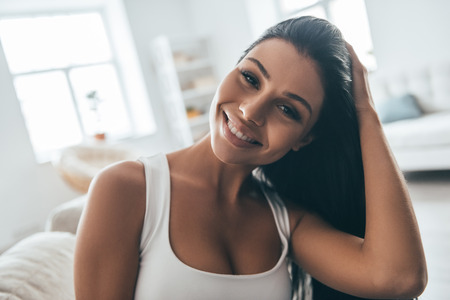 Enjoying free time at home. Portrait of attractive young woman adjusting her hair and looking at camera while sitting on the couch at homeの写真素材
