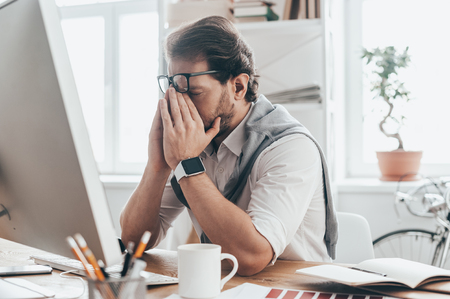 Exhausted day. Handsome young man with closed eyes touching face with hands while sitting on working place in creative officeの写真素材