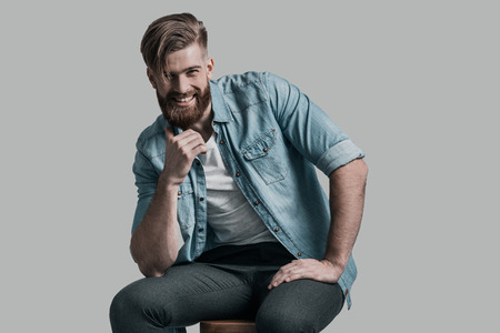 Too good to be real. Stylishly dressed young man smiling and holding hand on chin while sitting on chair against grey backgroundの写真素材