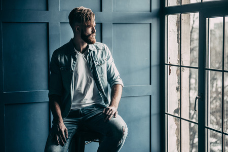 Young and confident. Handsome young man in light blue jeans shirt looking in window while sitting on chairの写真素材