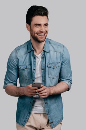 Receiving funny messages. Handsome smiling young man in blue jeans shirt holding smart phone and looking away while standing against grey backgroundの写真素材