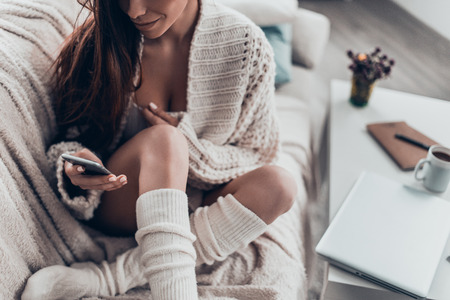 Texting to friends. Close-up of attractive young woman typing something using her smart phone while sitting on couch at homeの写真素材