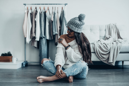 Lazy day at home. Beautiful young woman in casual wear and knit hat sitting on the floor and adjusting her sweater near her clothes hanging on the racksの写真素材