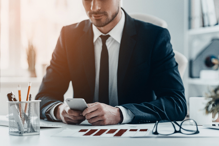 Using modern technologies at work. Close-up of young man in full suit holding smart phone while sitting at the office deskの写真素材