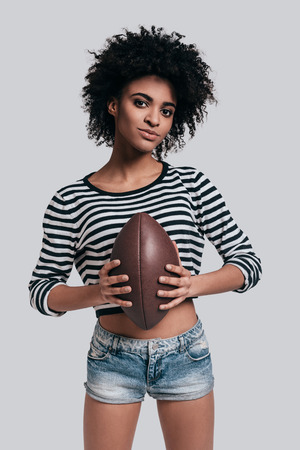 Attractive football player. Beautiful young African woman in casual wear carrying a ball and looking at camera while standing against grey backgroundの写真素材