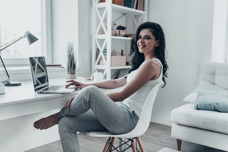 Surfing the net at home. Beautiful young woman using laptop and looking at camera with smile while sitting at the desk at homeの写真素材