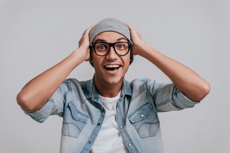 Unexpected news. Surprised young man in jeans shirt holding hands on head and looking at camera while standing against grey backgroundの写真素材