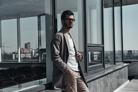 Young and successful.  Confident young man in eyewear keeping hand in pocket and looking away while leaning on the office building outdoorsの写真素材