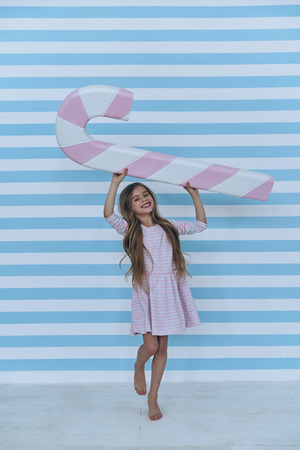 Happy to have candy. Lovely little girl looking at camera and smiling while holding a giant candy cane above her headの写真素材