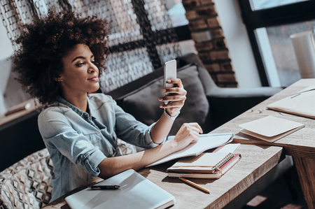 Answering important messages. Beautiful young African woman looking at smart phone and smiling while sitting at her working place in restaurantの写真素材