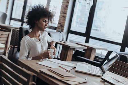 Enjoying fresh coffee at her favorite place. Attractive young African woman holding a cup and looking away with smile while sitting in restaurantの写真素材